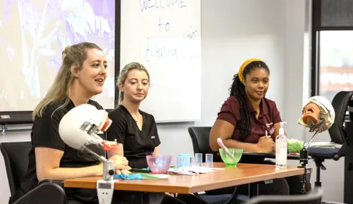 Three young women in Germanna attire sit at a table with medical training equipment giving a demonstration