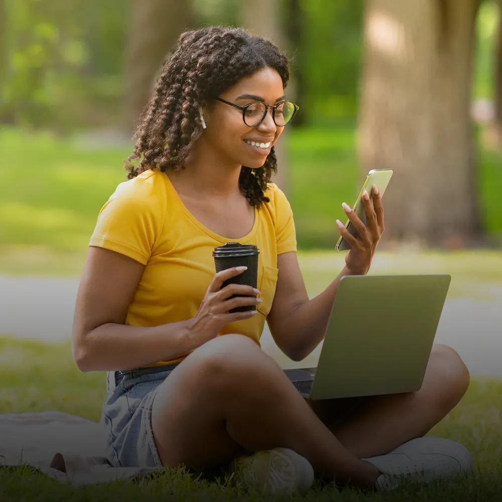 A woman relaxes outside on the grass while applying for summer classes on her phone